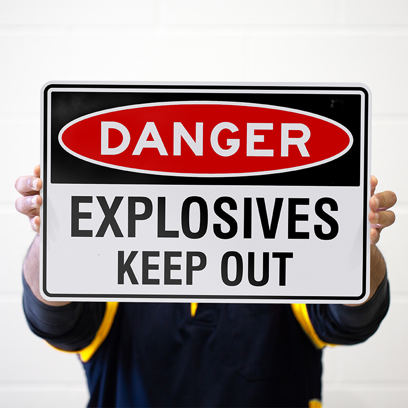 A person holding a sign that reads 'DANGER EXPLOSIVES KEEP OUT' in bold black and white text against a red background.