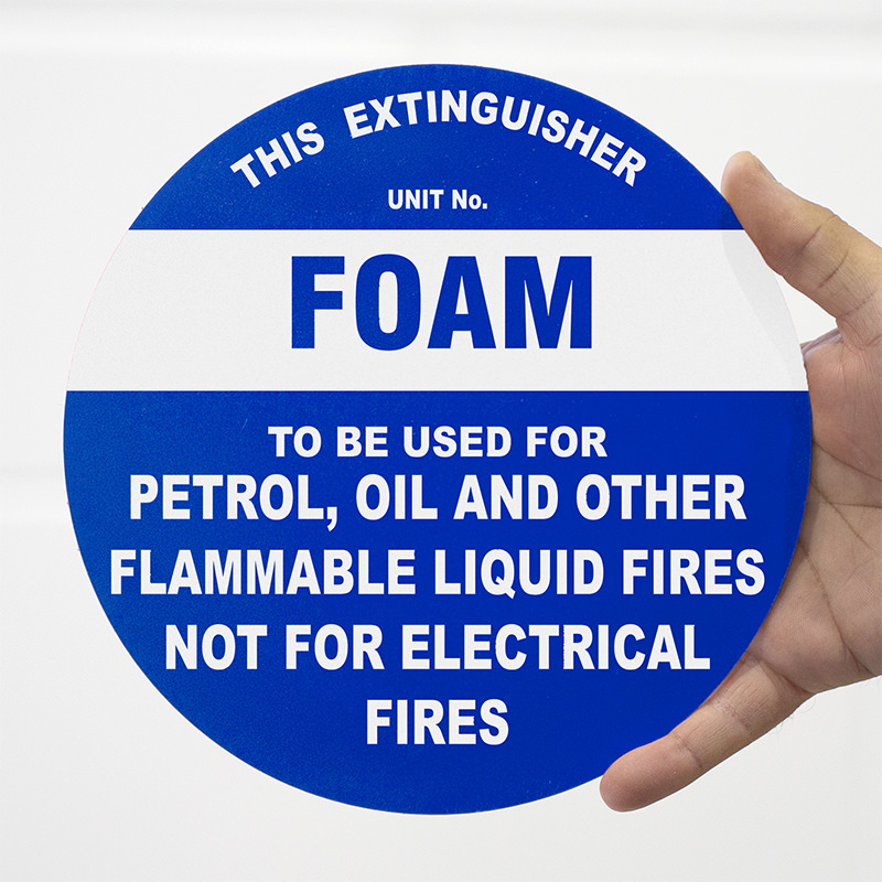 A person holding a circular blue sign that reads 'FOAM' in large white letters, with the text 'THIS EXTINGUISHER UNIT No. TO BE USED FOR PETROL, OIL AND OTHER FLAMMABLE LIQUID FIRES NOT FOR ELECTRICAL FIRES' underneath.