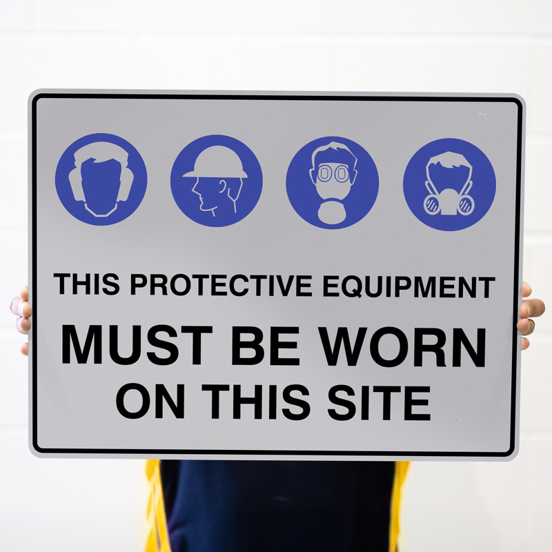 A person holding a large sign that features icons of protective equipment, including ear protection, a hard hat, a face mask, and a respirator, with the text 'THIS PROTECTIVE EQUIPMENT MUST BE WORN ON THIS SITE' prominently displayed in bold letters.