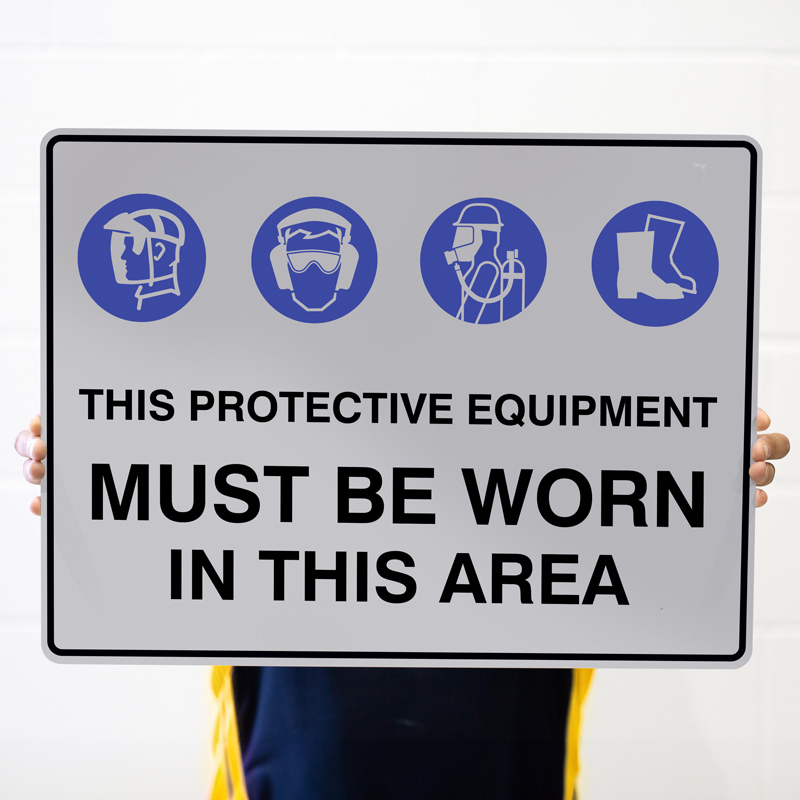 A person holding a sign that indicates protective equipment must be worn in the area, featuring icons for a safety helmet, goggles with ear protection, a respirator, and safety boots.