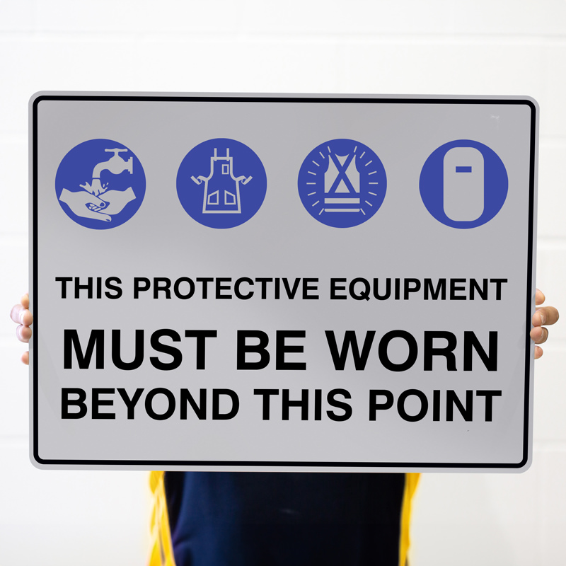 A person holding a safety sign that reads 'THIS PROTECTIVE EQUIPMENT MUST BE WORN BEYOND THIS POINT,' featuring icons of protective gear including gloves, a protective apron, a safety helmet, and a face shield.