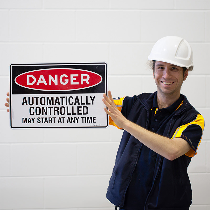Smiling worker in a hard hat holding a danger sign that reads Automatically controlled may start at any time